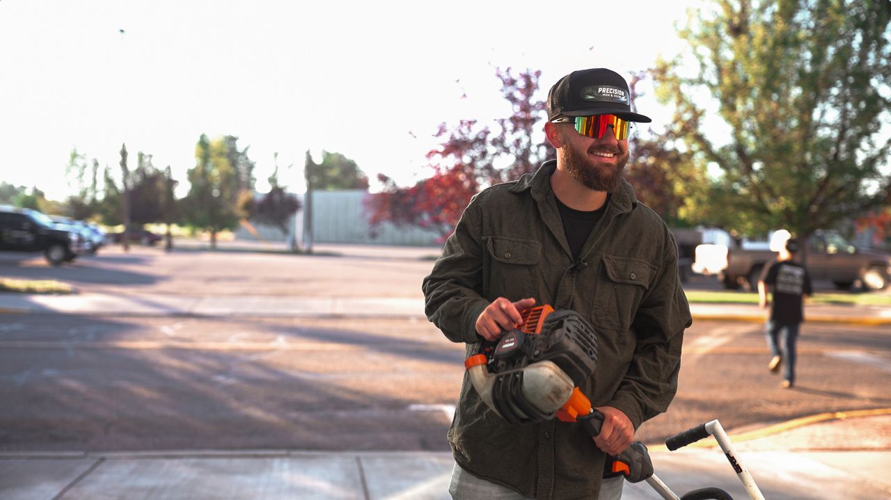 Smiling crew member in a Precision cap and sunglasses holding a string trimmer in a parking lot
